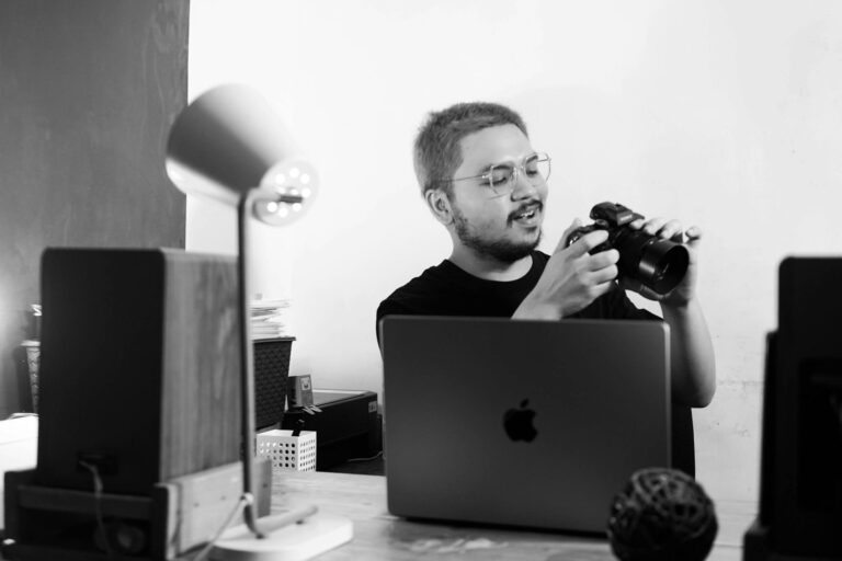 Black and white photo of a man reviewing a camera at his desk in a modern office environment.